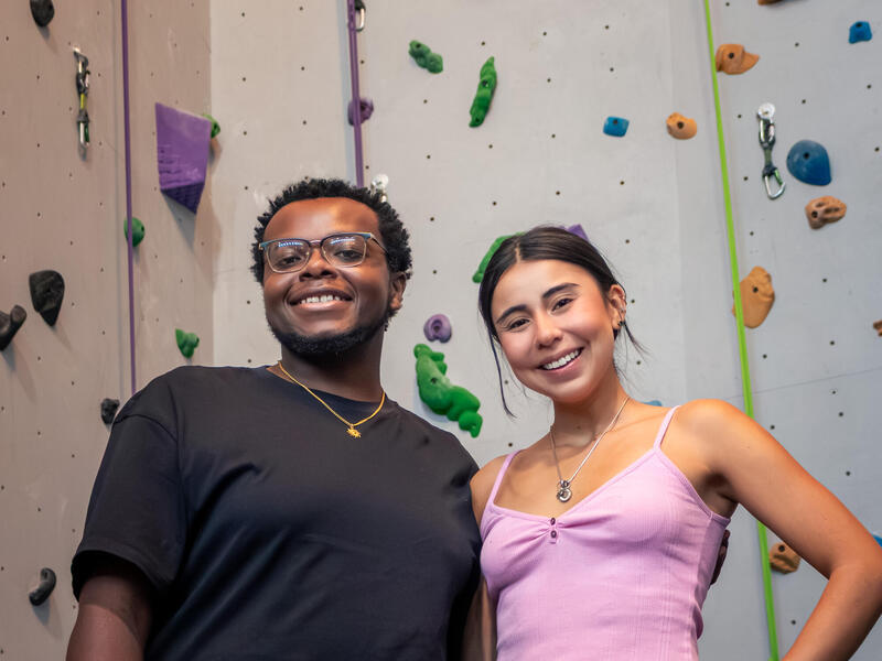 Two people stand and smile in front of an indoor rock climbing wall.