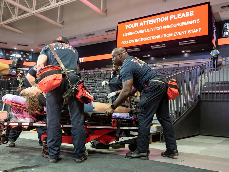 A photo of three EMS workers standing around a woman who is on a stretcher in a basketball stadium. 
