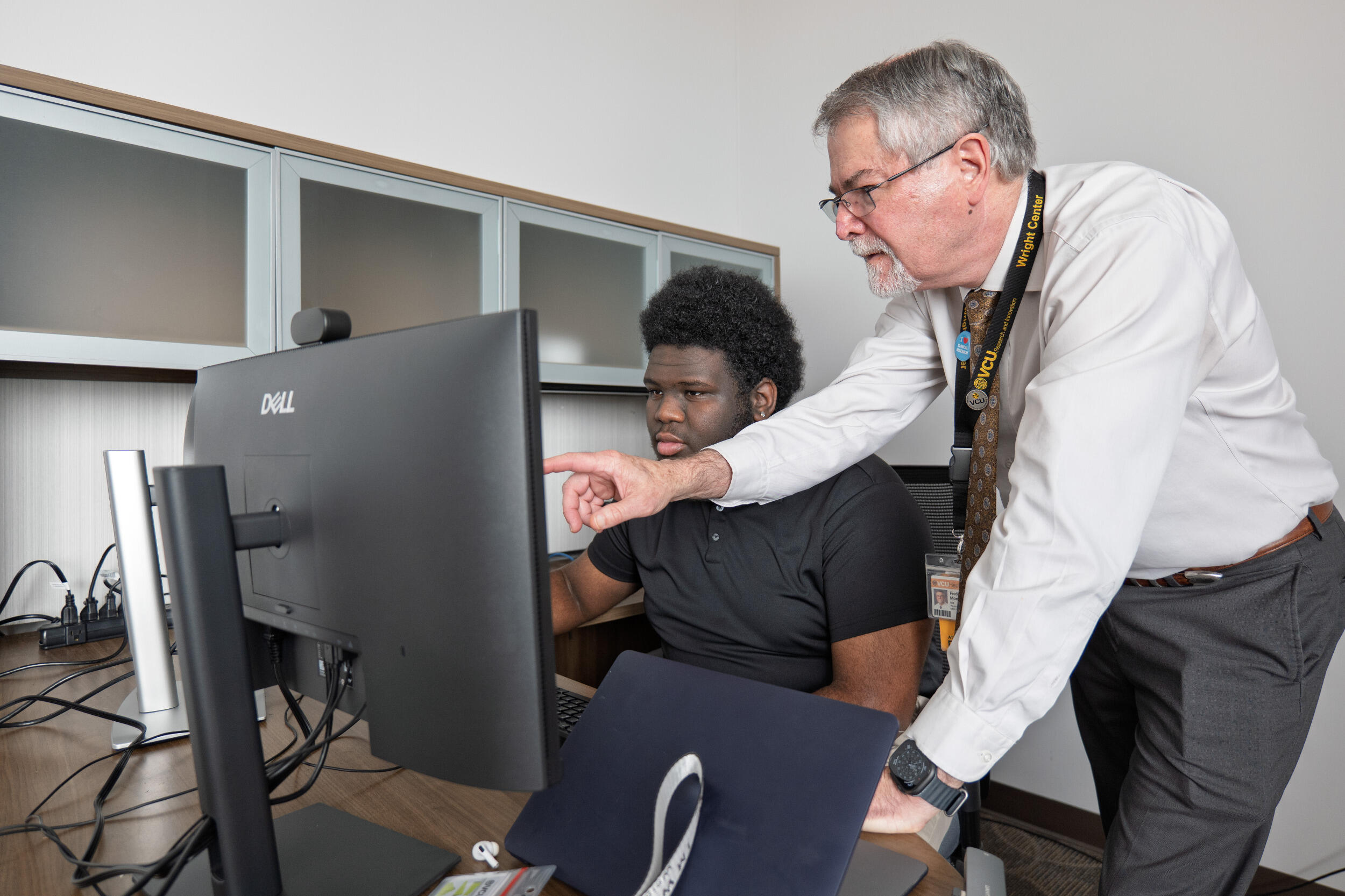 A man sits at a computer looking at the screen while another man stands and points something out on the screen.