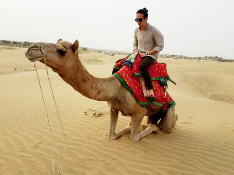 A man sits on a camel with a red saddle in a desert.