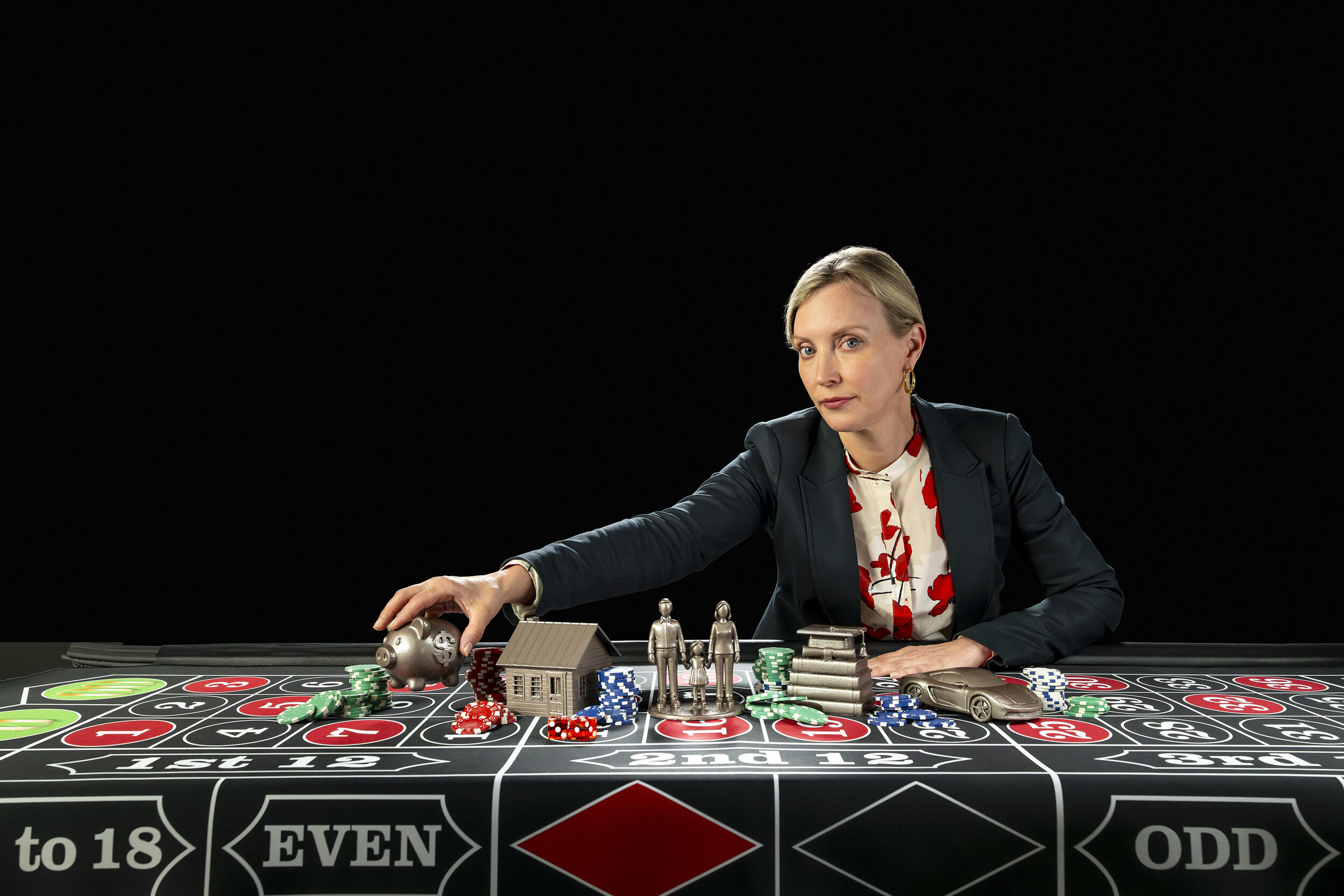 A photo of a woman sitting at a table covered in gambling chips and bronze statues of two people, a house, a pile of books with a graduation cap on top, a piggy bank, and a car. 