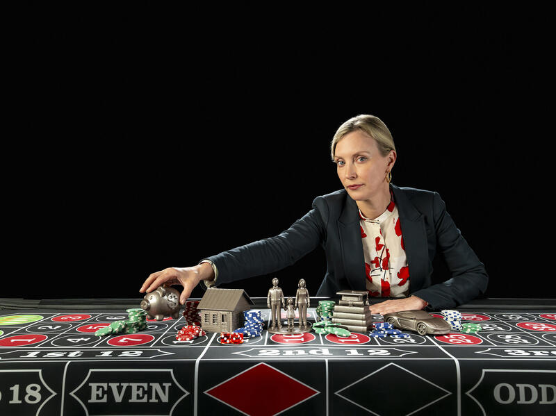 A photo of a woman sitting at a table covered in gambling chips and bronze statues of two people, a house, a pile of books with a graduation cap on top, a piggy bank, and a car. 