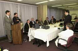 Group of people, two of whom are standing at a lectern and others are seated around a table.