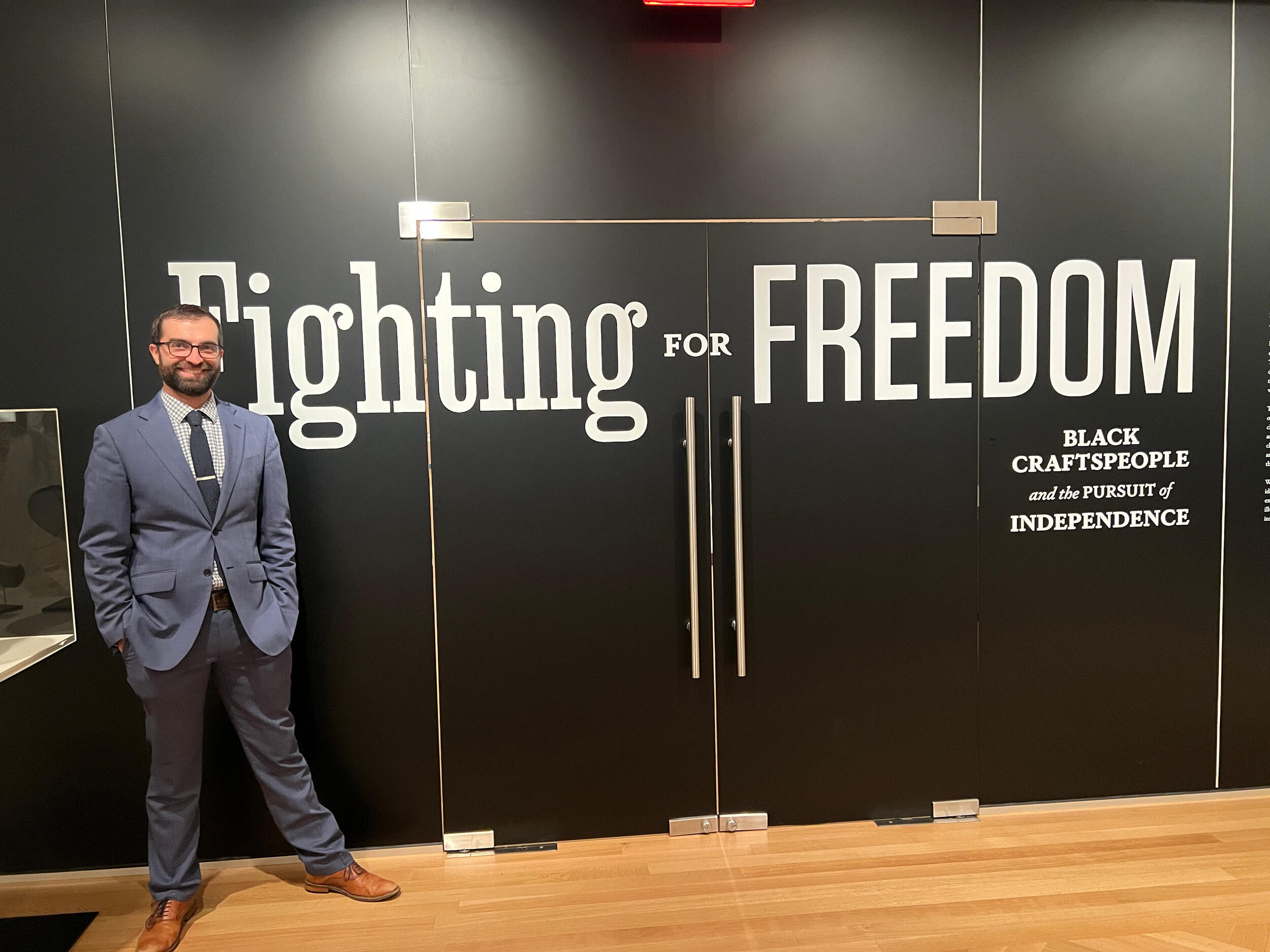 A photo of a man standing next to a black wall. White letters on the wall spell out \"Fighting FOR FREEDOM\" \"BLACK CRAFTSPEOPLE and the PURSUIT of INDEPENDENCE\"