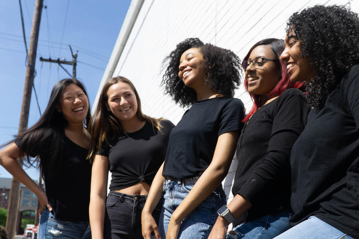 Diverse group of 5 girls, all in black shirts, stand together smiling. 