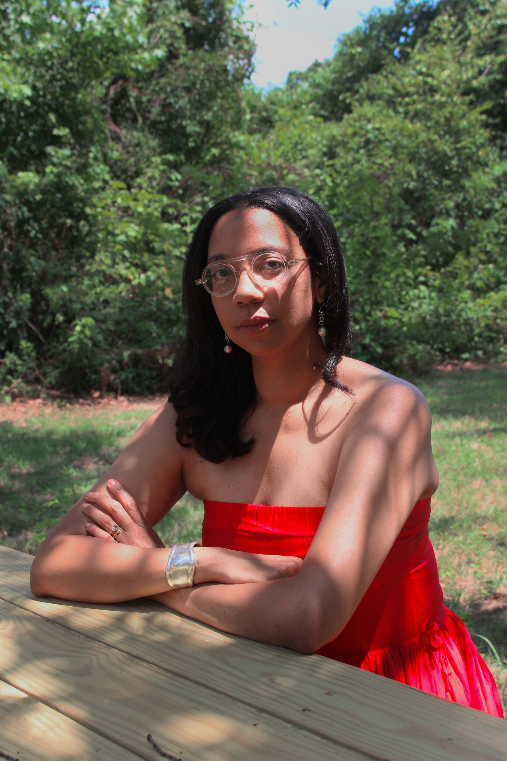 A photo of a woman sitting at a wooden table outside. There are trees and grass behind her. 