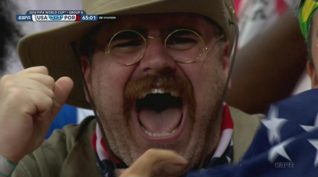 A man in a mustache, glasses and a hat cheers.