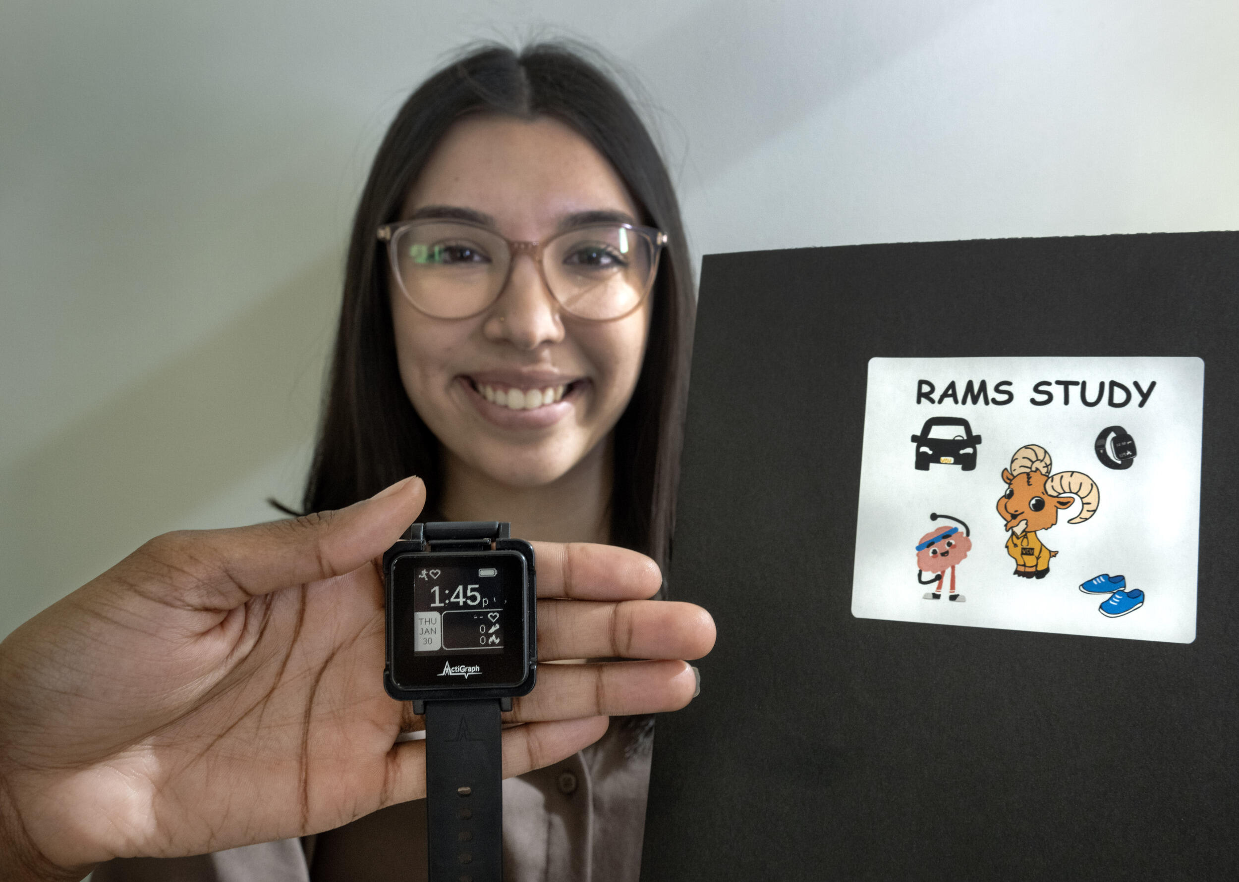 A photo of a woman smiling and holding up a folder that says \"RAMS SUTDY\" in comic sans font. On the folder are illustrations of a car, a cresent moon, a ram that is smiling, tennis shoes, and a brain with arms, legs, and a face wearing a sweat headband and sneakers stretching it's right arm over top itself. Another person's hand is holding a smart watch. 