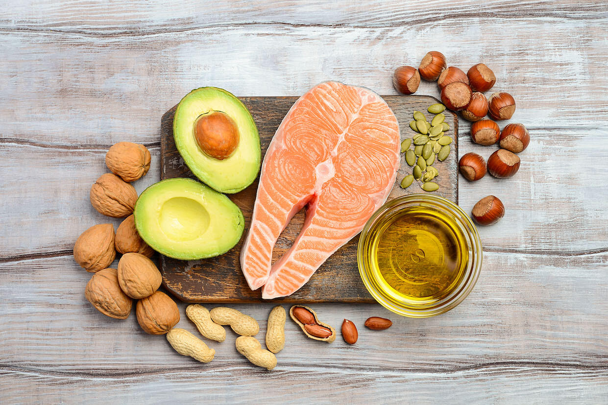 A cutting board with a cut of salmon, a split avocado, seeds and a cup of olive oil on it with hazelnuts, peanuts and walnuts around its sides.