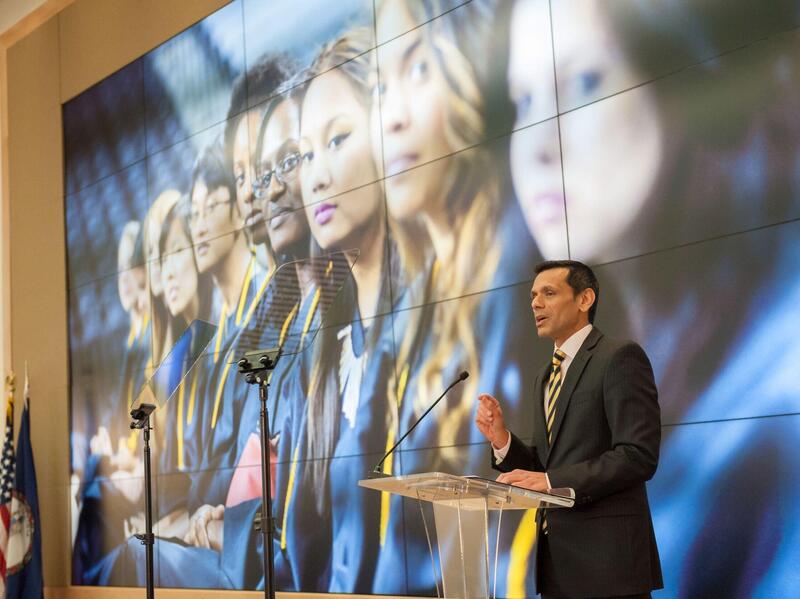 Photo of Michael Rao speaking at a podium with a large image behind him of a group of people looking toward the camera.