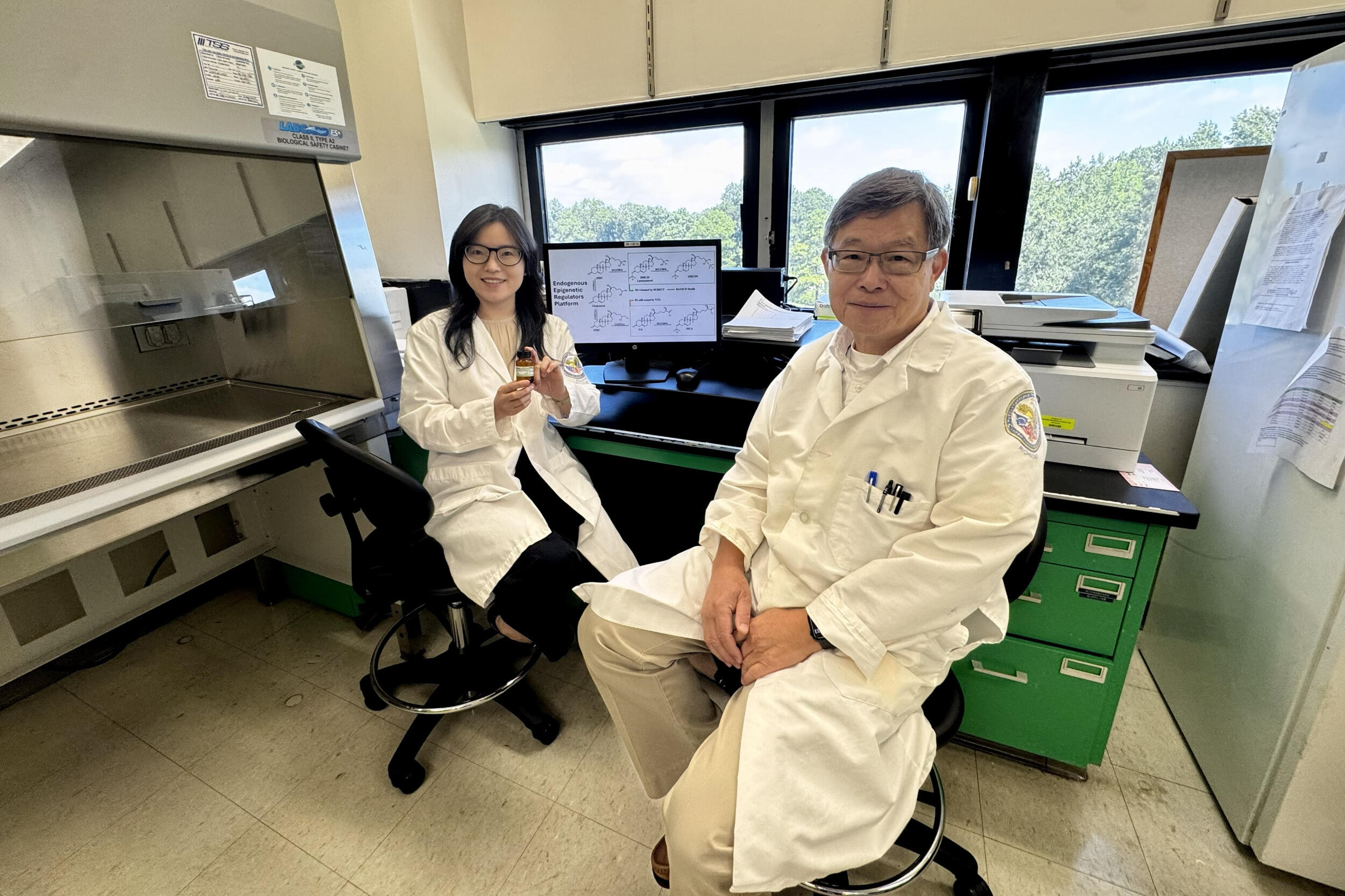 A woman and man in lab coats sit on chairs. The woman holds up a bottle.