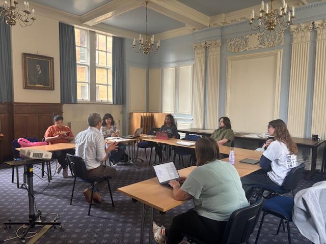 A photo of students sitting in tables positioned around a professor who is sitting. 