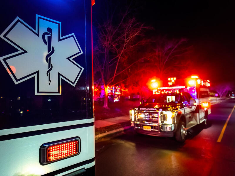 A photo of an ambulance up close and one parked behind it. The ambulance in the back has its emergency lights on. 