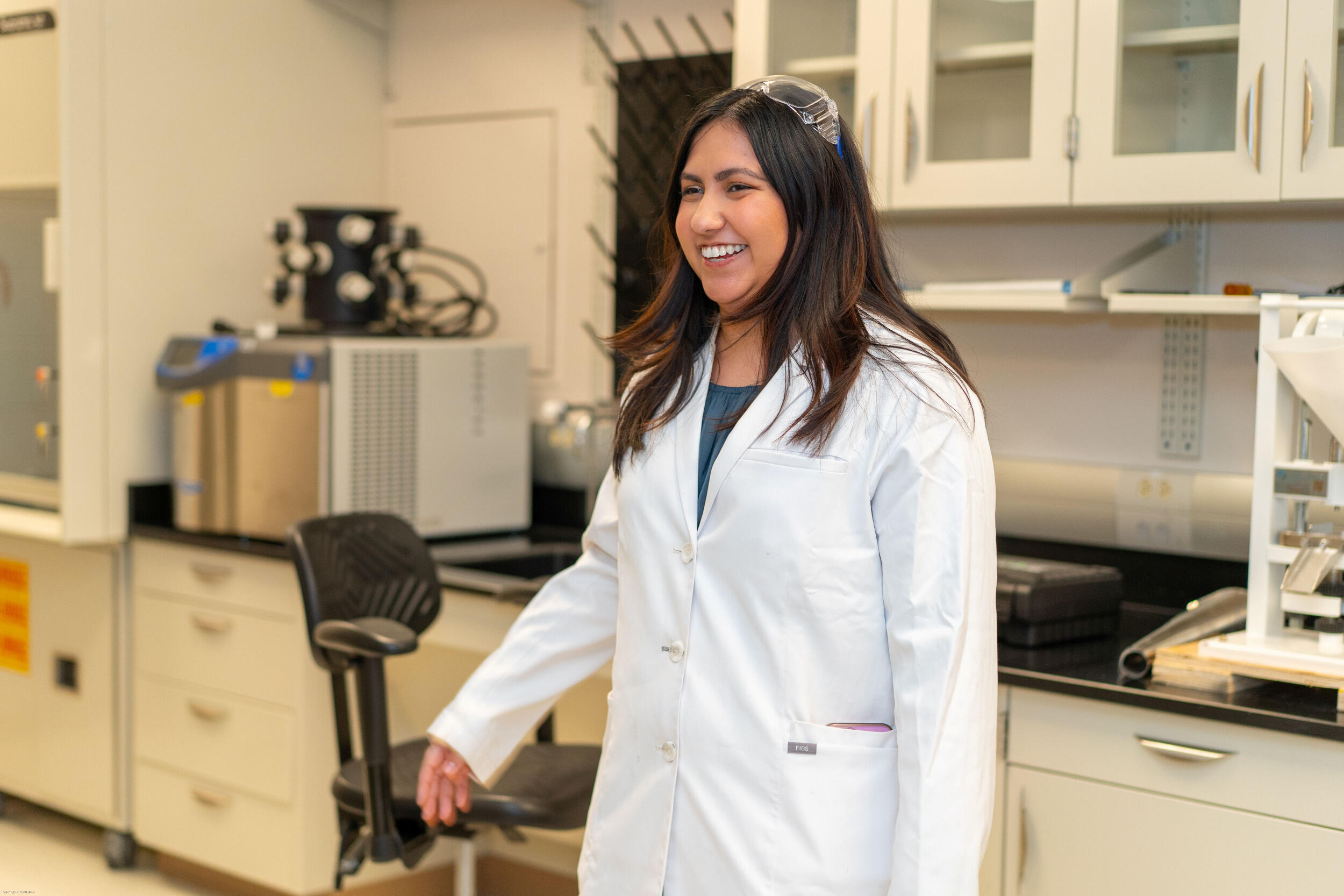 A woman in a lab coat and safety goggles smiles in a lab setting.