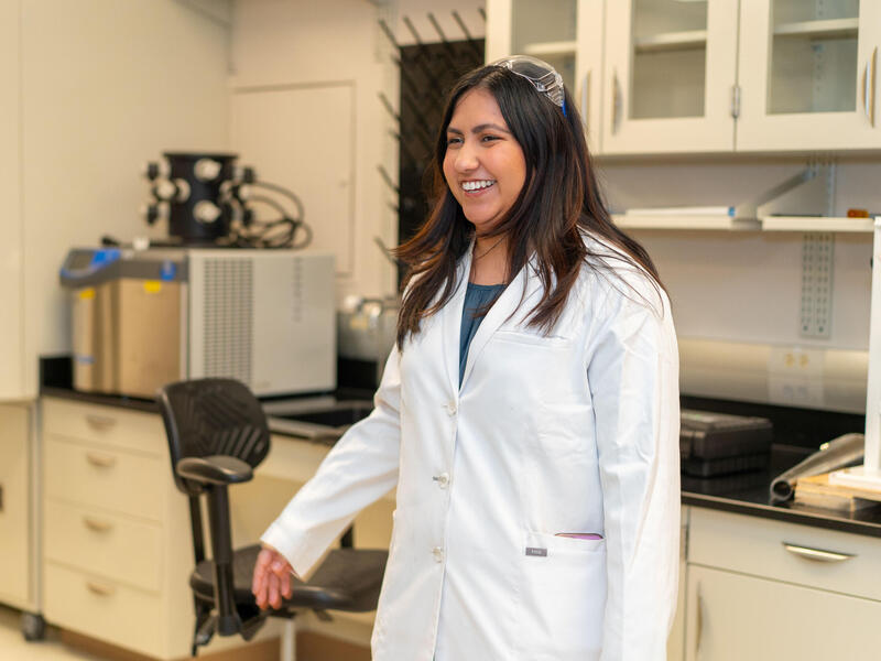 A woman in a lab coat and safety goggles smiles in a lab setting.