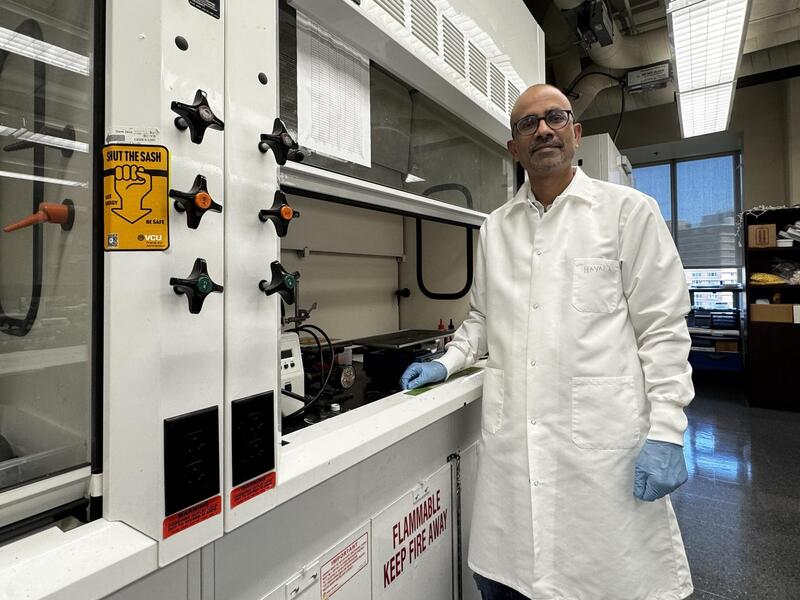 A photo of a man in a white lap coat, blue latex gloves, and safety glasses standing next to a fume hood in a science lab. 