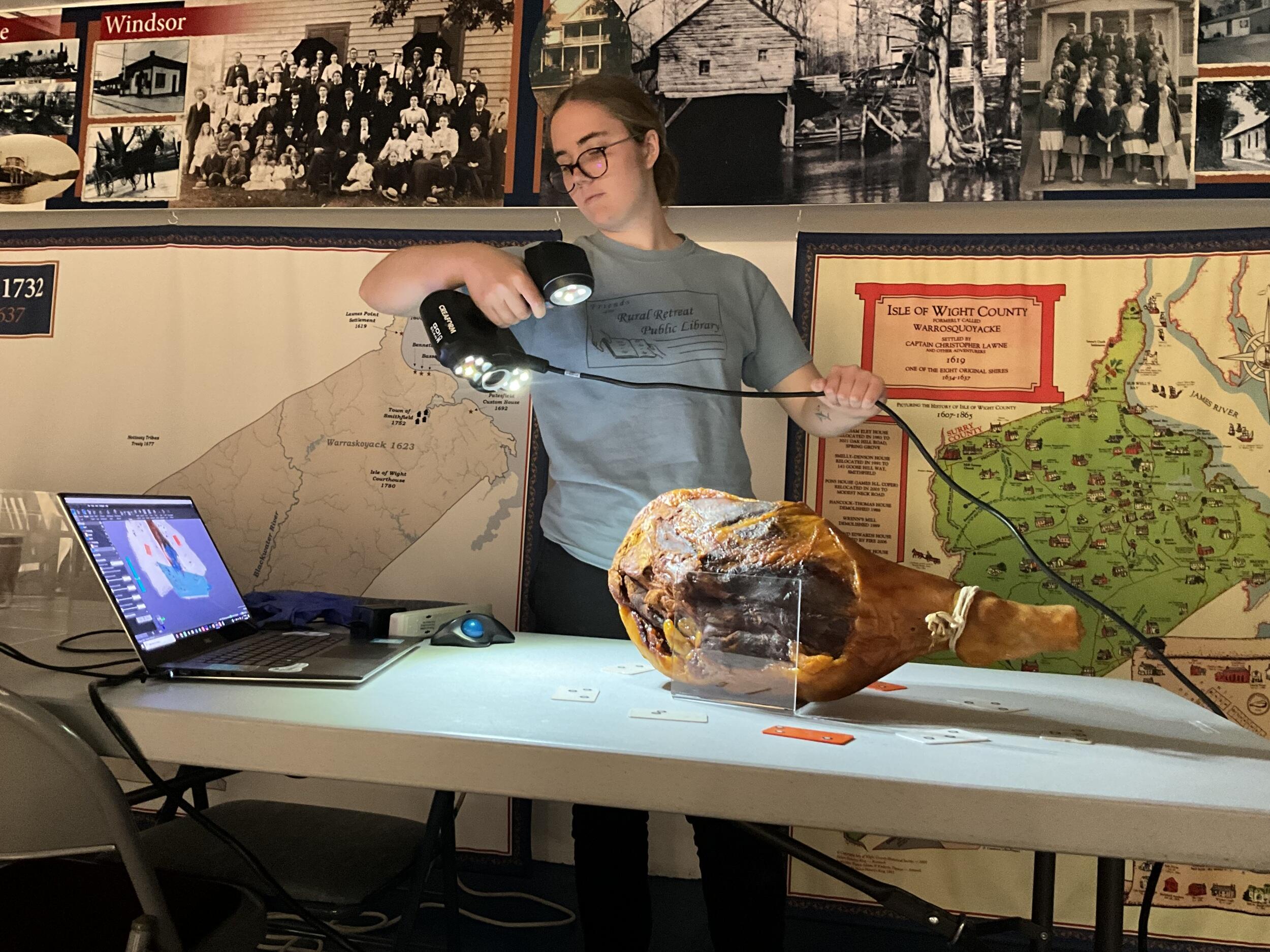 A photo of a woman shining a light on a ham. The ham is sitting on the table with a laptop. Behind the woman and the table are old black and white photos of people, and a geographic map. 