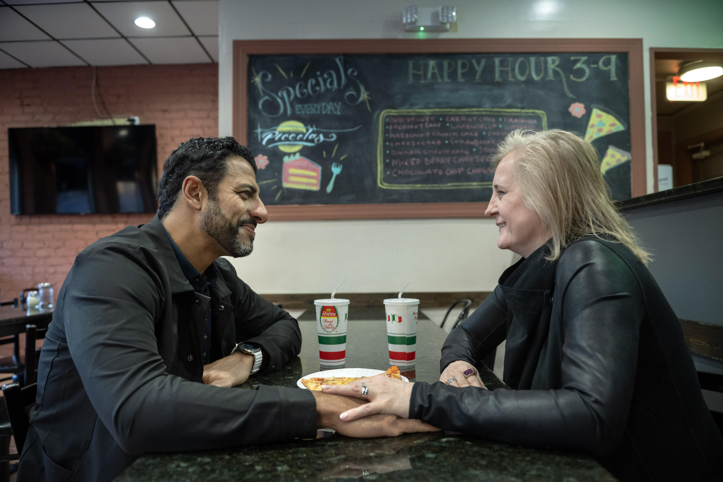 A photo of a man and a woman sitting across from each other at a table. They are looking at each other lovingly and the woman has her hand on one of the man's. 