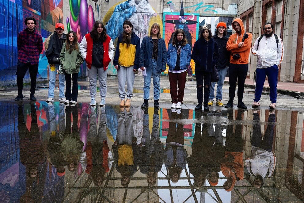 A group photo of ten people standing in front of a puddle with the reflection of the puddle being the ten people standing. 