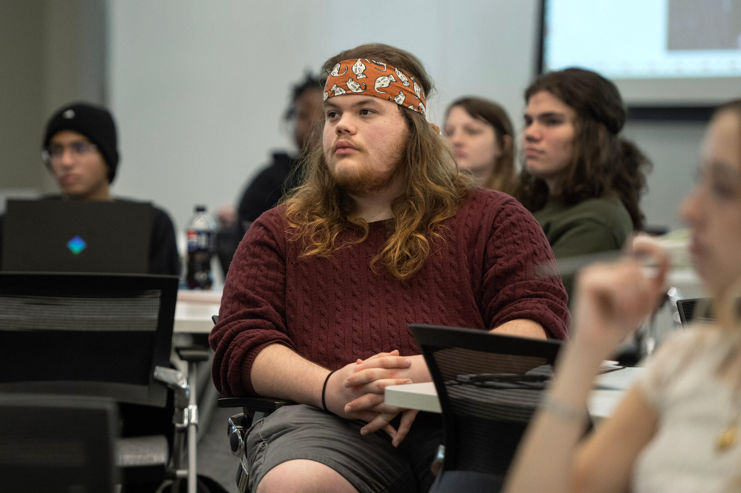 A person in a sweater and bandanna sits at a table and looks toward the front of the room.