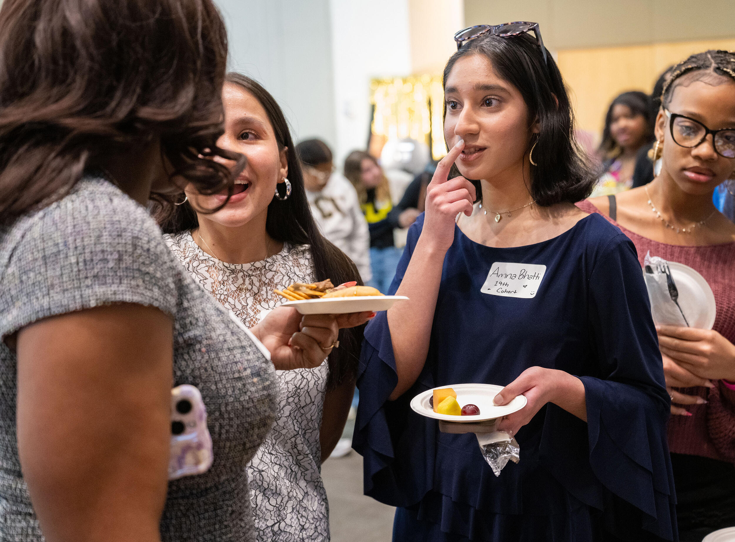 A photo of four women standing. Two are holding plates with food and one is looking at their phone. 