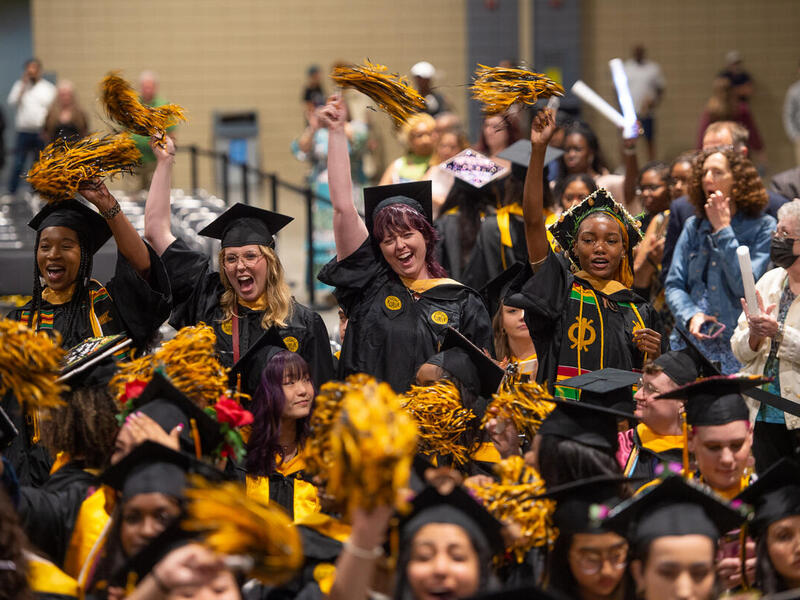 A photo of a crowd of people wearing graduation caps and gowns cheering and waving pompoms in the air. 