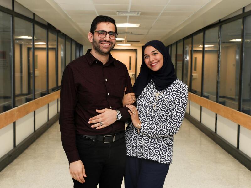 A photo of a mand and a woman standing and smiling. They are holding arms and standing in a hallway. 