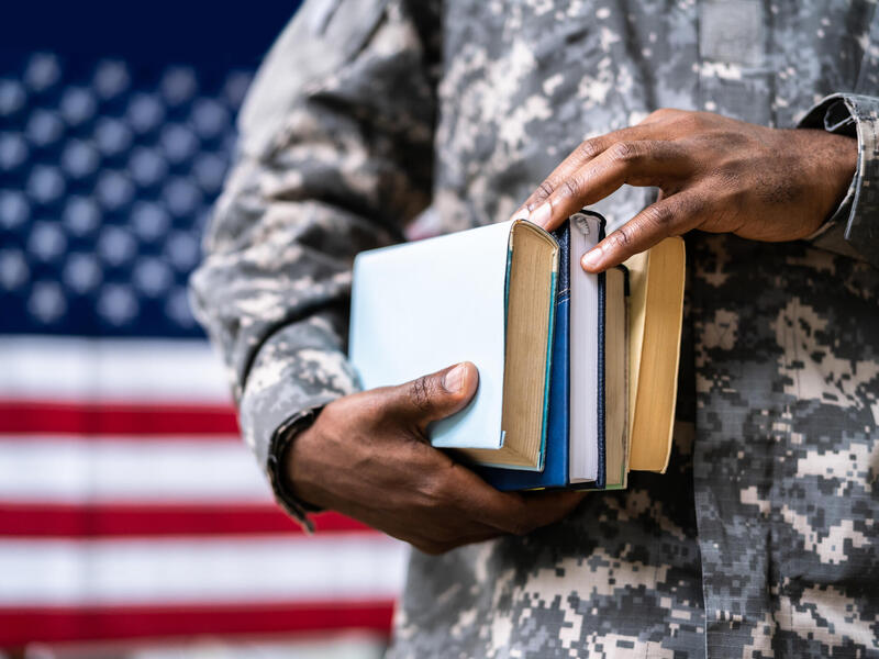 A photo of a person in a military uniform from the shoulders down. They are holding a stack of four books and standing in front of an American flag. 