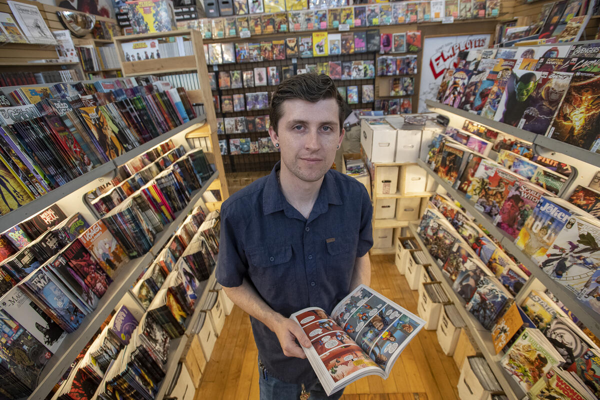 A man standing in the middle of a comic shop holding an open comic book. All around him are shelves lined with comic books and graphic novels. 