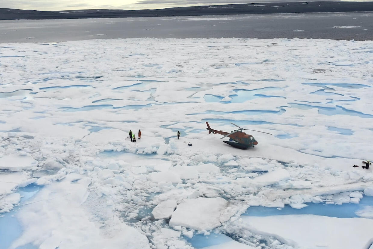 A group of researchers stands on a frozen block of ice in the Arctic. A helicopter sits nearby with various pieces of equipment.