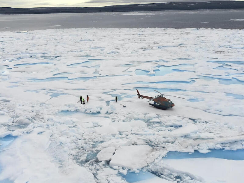A group of researchers stands on a frozen block of ice in the Arctic. A helicopter sits nearby with various pieces of equipment.