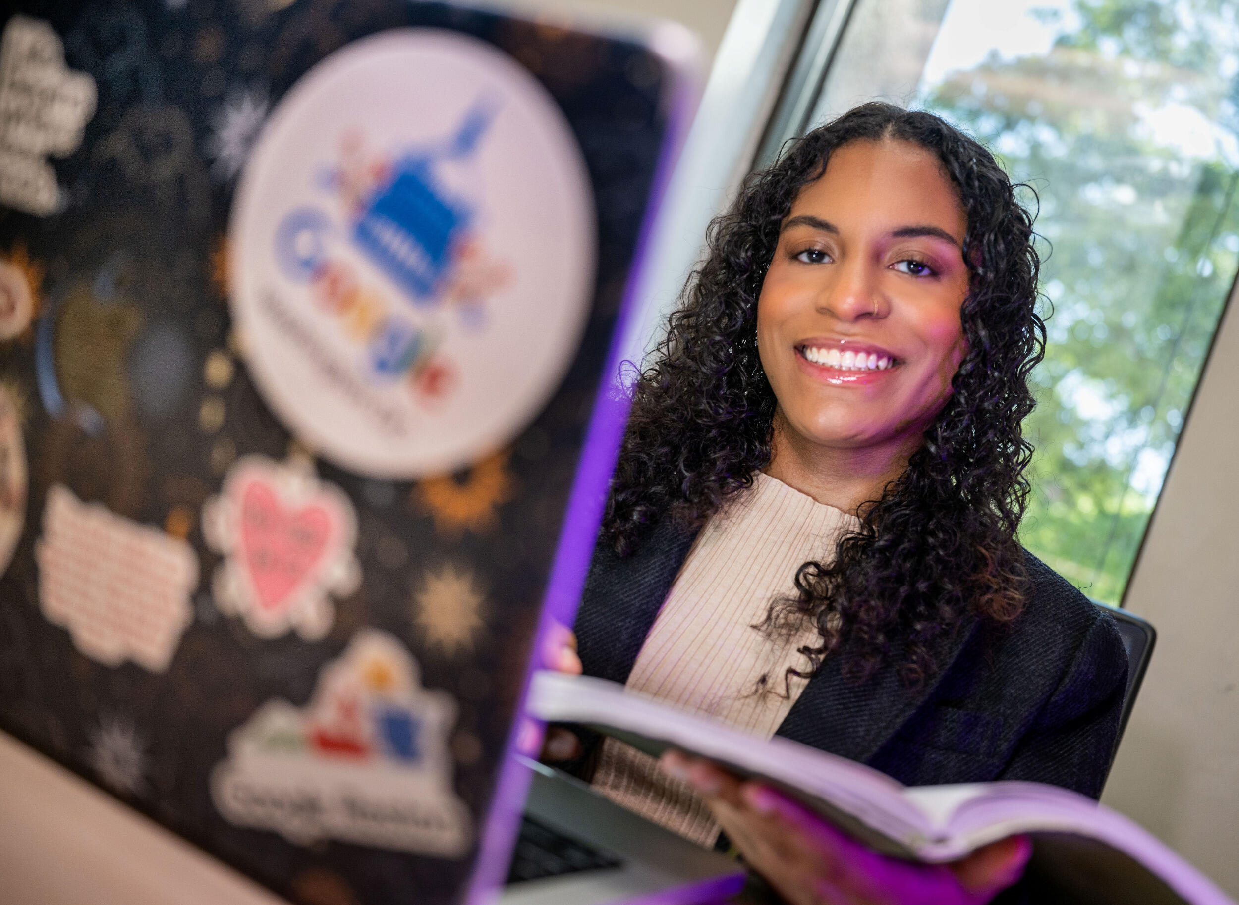 A woman holds an open book in front of an open laptop.