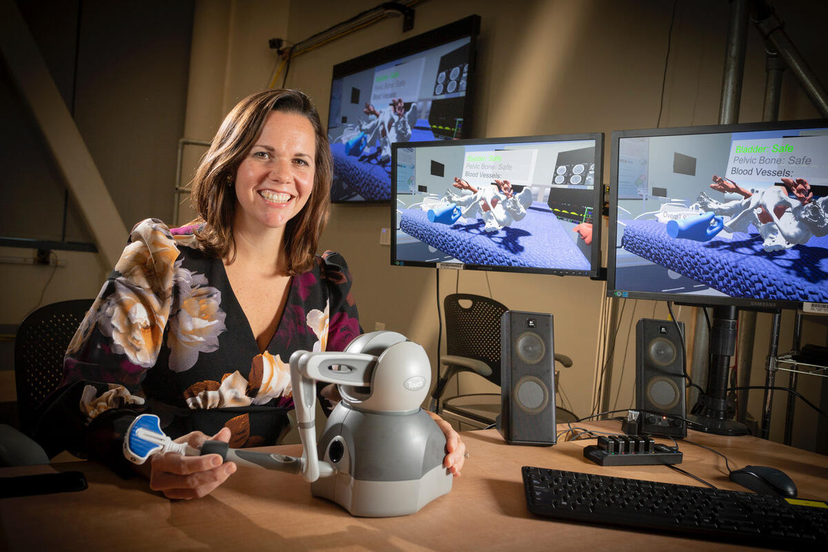 A woman holding a piece of robotics equipment sitting next to three computer screens with a 3D scenario on them