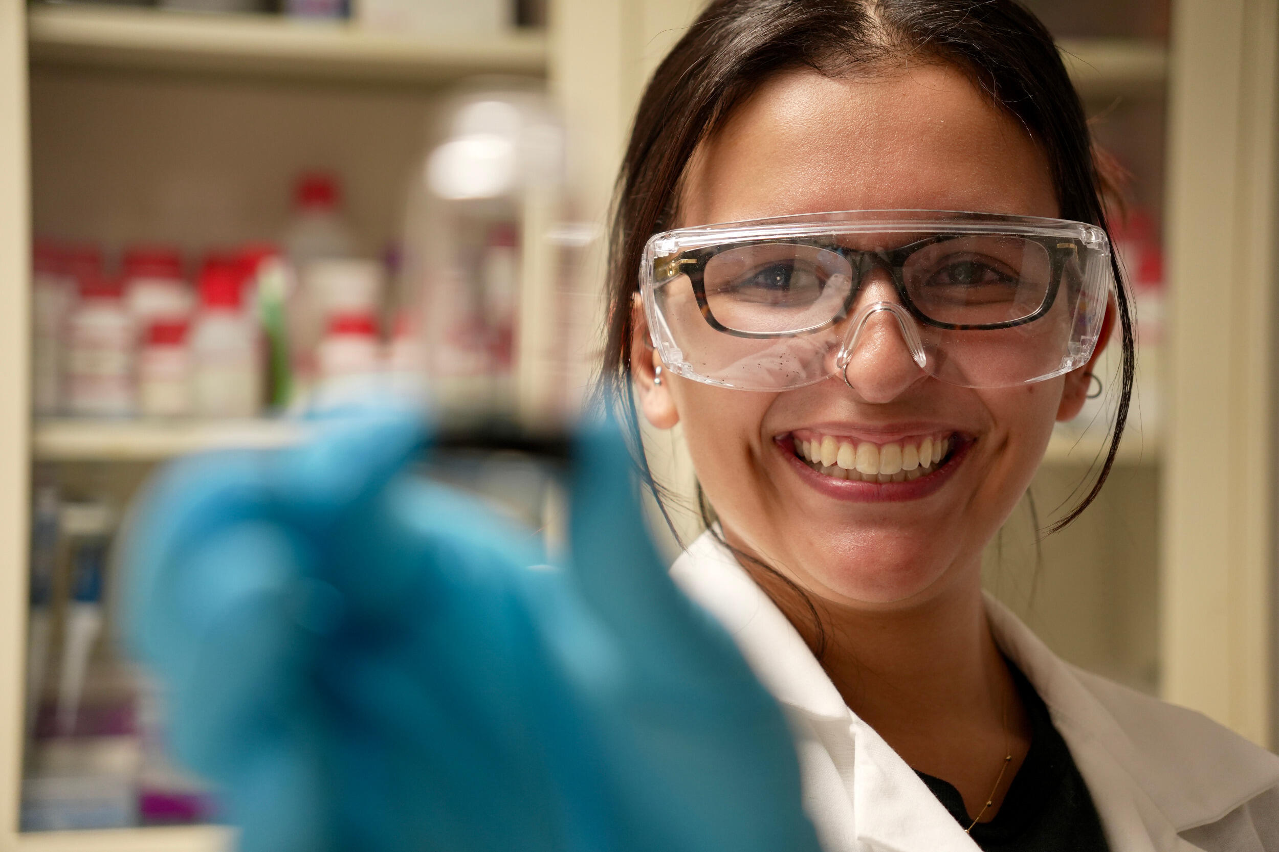 A woman in safety goggles and blue gloves smiles and holds up something to the camera.