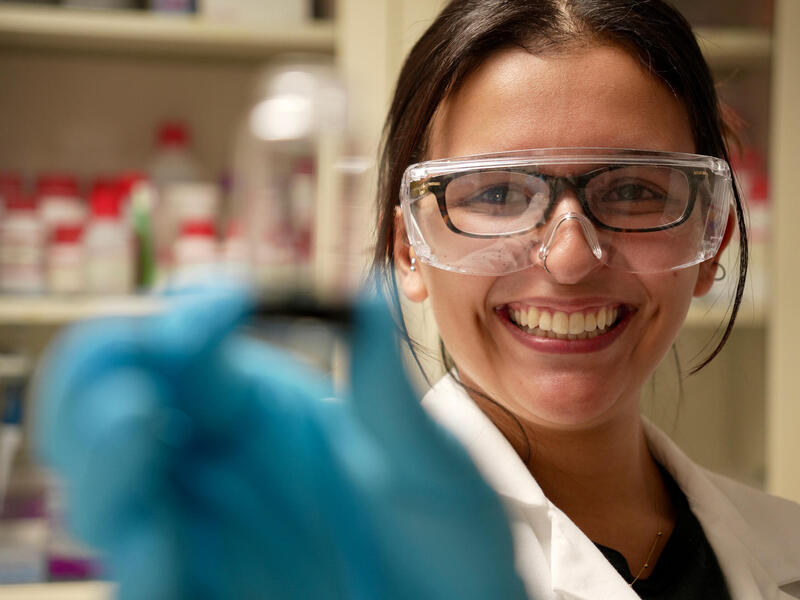 A woman in safety goggles and blue gloves smiles and holds up something to the camera.