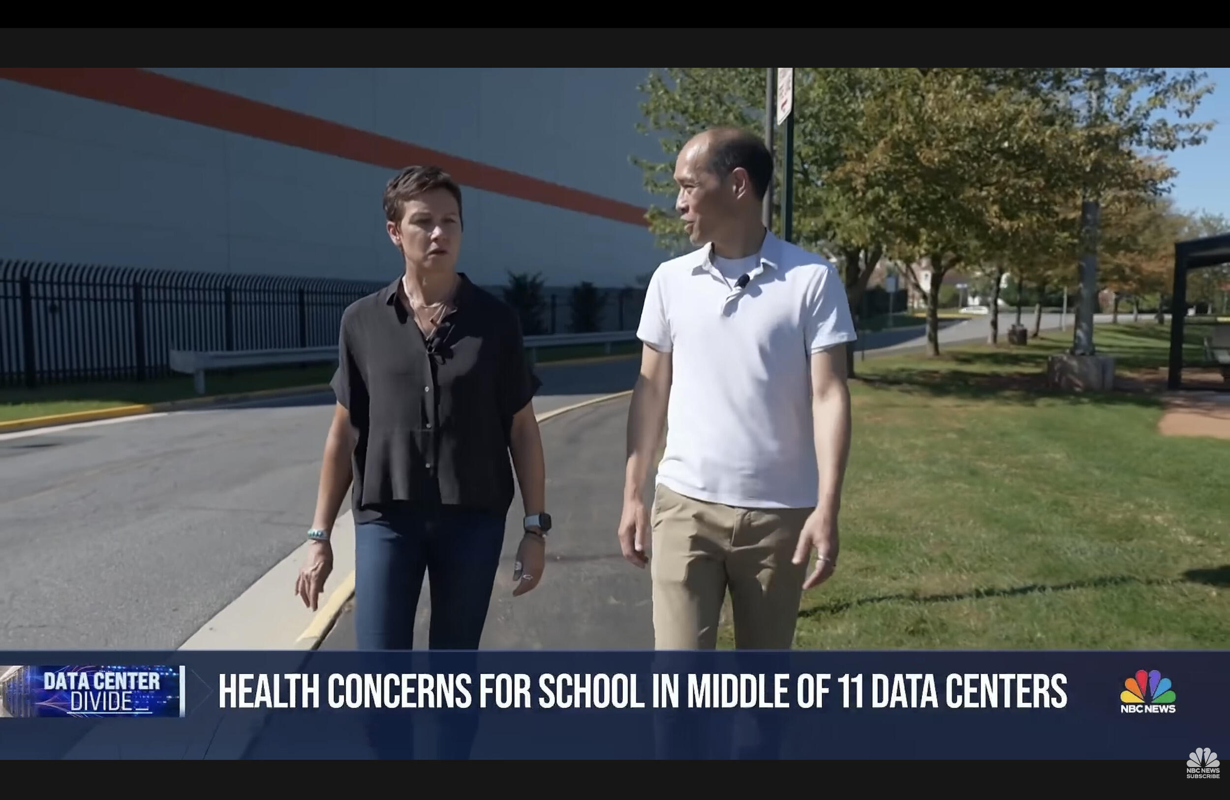 A screenshot of an NBC news boradcast. A mand and a woman are walking past a large building, and the man on the right is talking, On the bottom of the screen white text reads \"HEALTH CONCERNS FOR SCHOOL IN MIDDLE OF 11 DATA CENTERS.\"