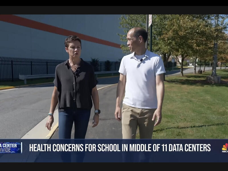 A screenshot of an NBC news boradcast. A mand and a woman are walking past a large building, and the man on the right is talking, On the bottom of the screen white text reads \"HEALTH CONCERNS FOR SCHOOL IN MIDDLE OF 11 DATA CENTERS.\"