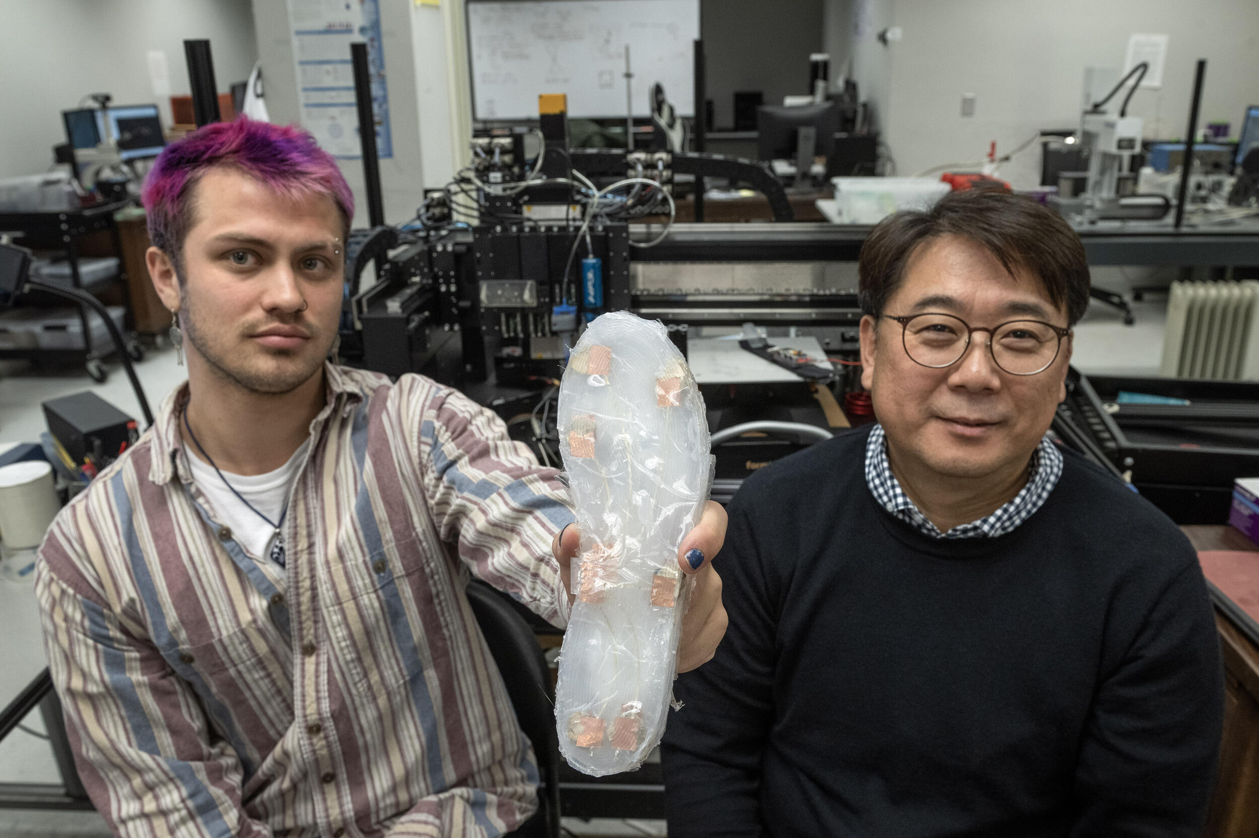 A photo of a two men sitting next to each other in a lab room. The man on the left is holding up opaque shoe sole. 