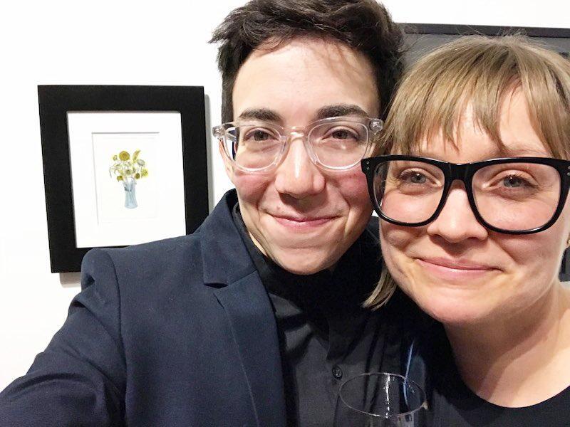 Two people wearing glasses smiling next to a framed picture of a flower vase.