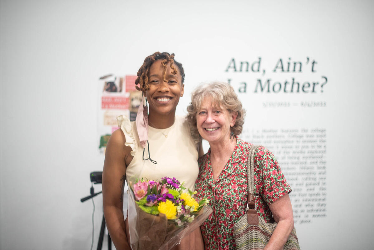 Two women standing close together in front of a wall with the words \"And, Ain't I a Mother?\" written on it. 