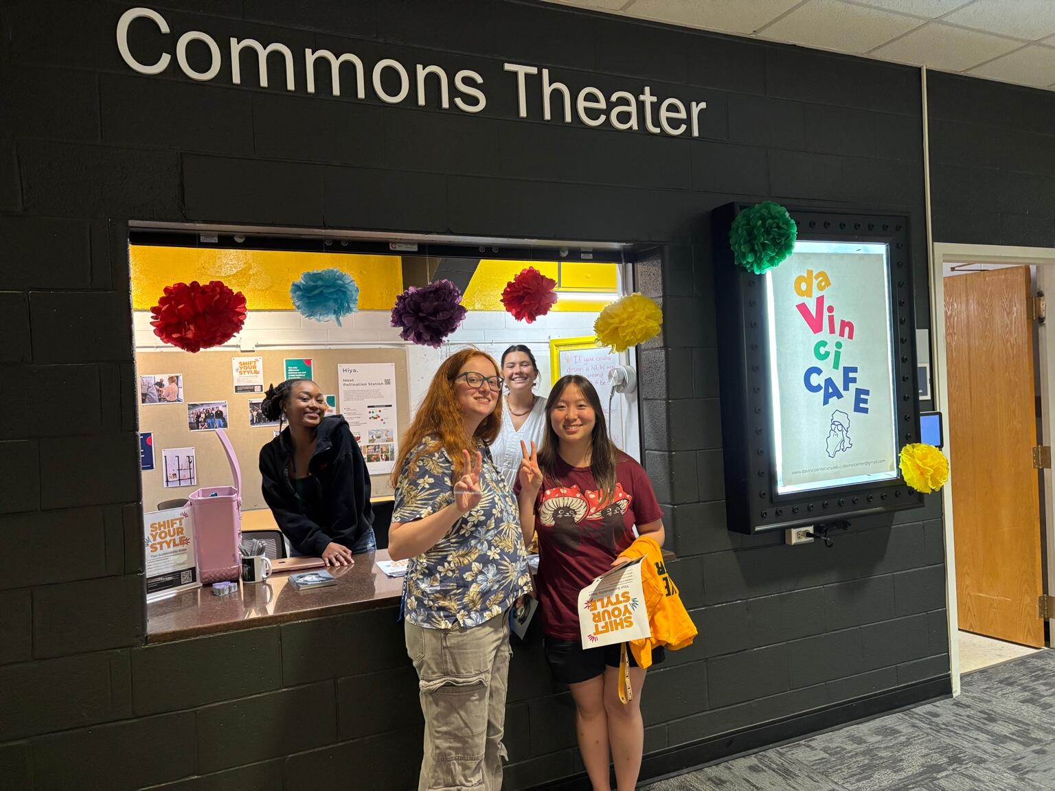 A photo of two women standing in front of a window that has a food counter. Behind the counter are two other women smiling. To the right of the window is a sign that says \"da Vinci Cafe\" 