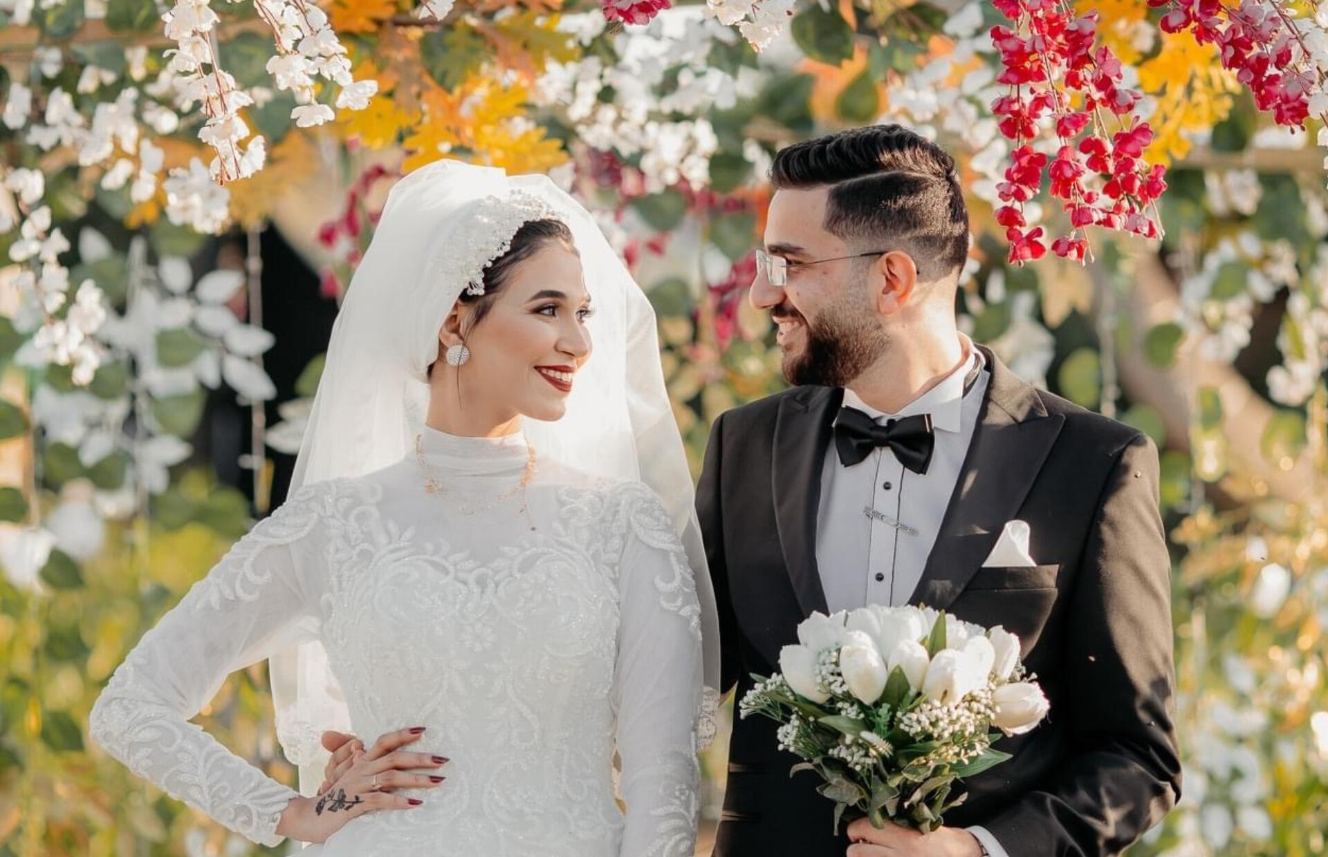 A photo of a man and a woman surrounded by flowers. The woman is in a bridal dress and viel, while the man is wearing a tux with a bowtie. 