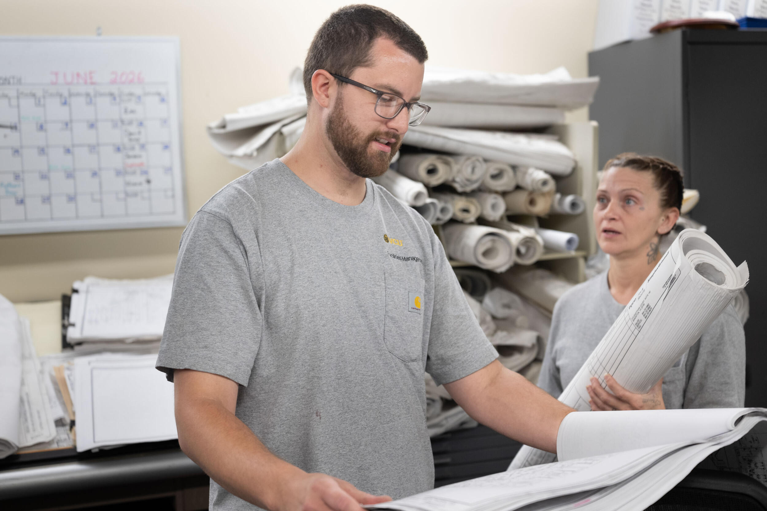 A photo of a man looking at a large sheet of paper. To his left, a woman is holding a large rolled up tube of paper. 