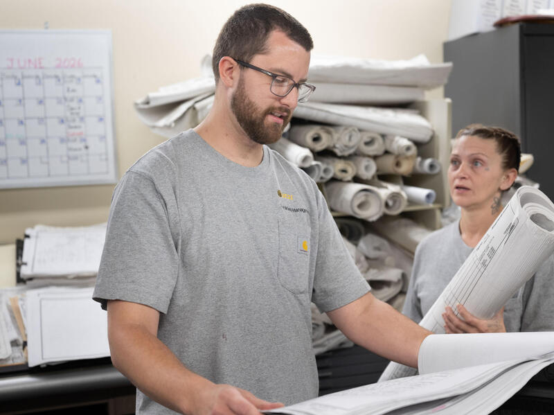 A photo of a man looking at a large sheet of paper. To his left, a woman is holding a large rolled up tube of paper. 