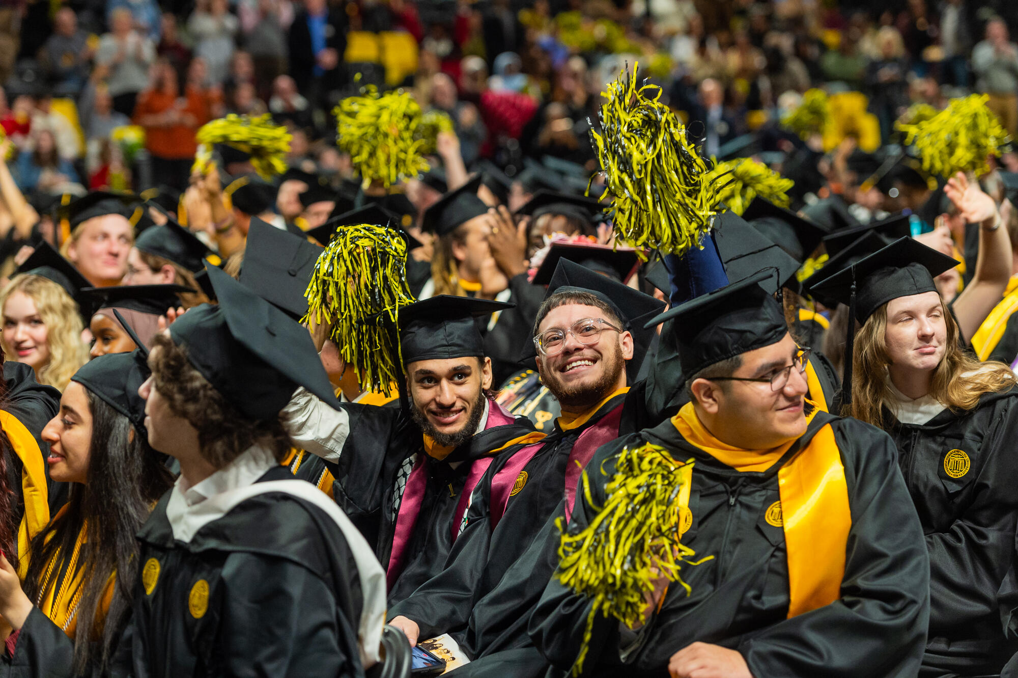 A photo of a crowd of graduates wearing graduation caps and gown.s Some of the people in the crowd are waving yellow and black pompoms. 