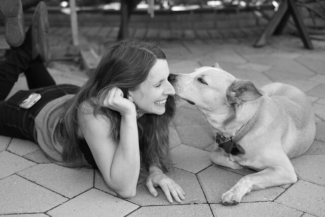 A photo of a woman laying on her stomach on the ground next to a dog who is sniffing her face while she smiles. 
