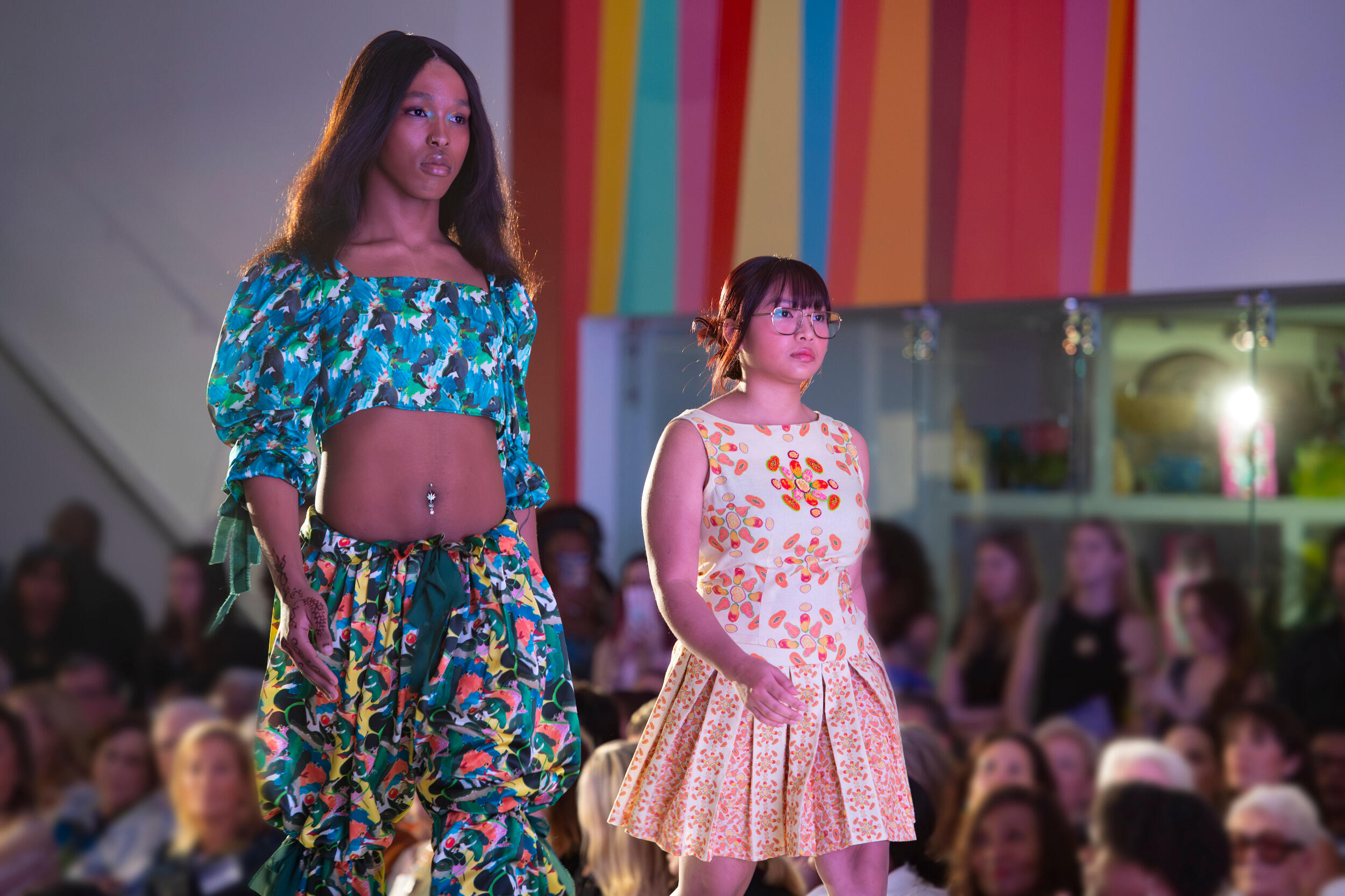 Two women in dresses walk in a fashion show in front of an audience.