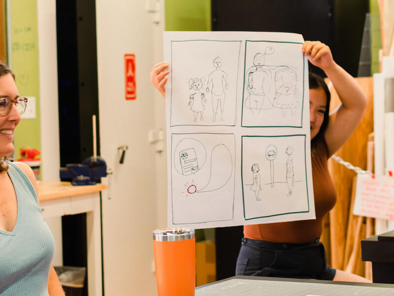 A photo of two mone. The woman on the right is holding up a piece of paper that has four children's illustrations on it. 