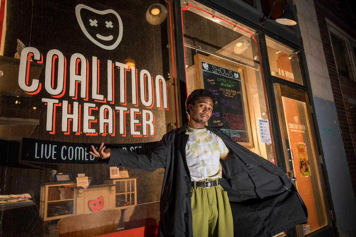 A person wearing a long black coat standing with their left arm extended and their right arm holding the coat open. They are standing in front of a window with the words \"COALITION THEATER\" painted in white letters. 