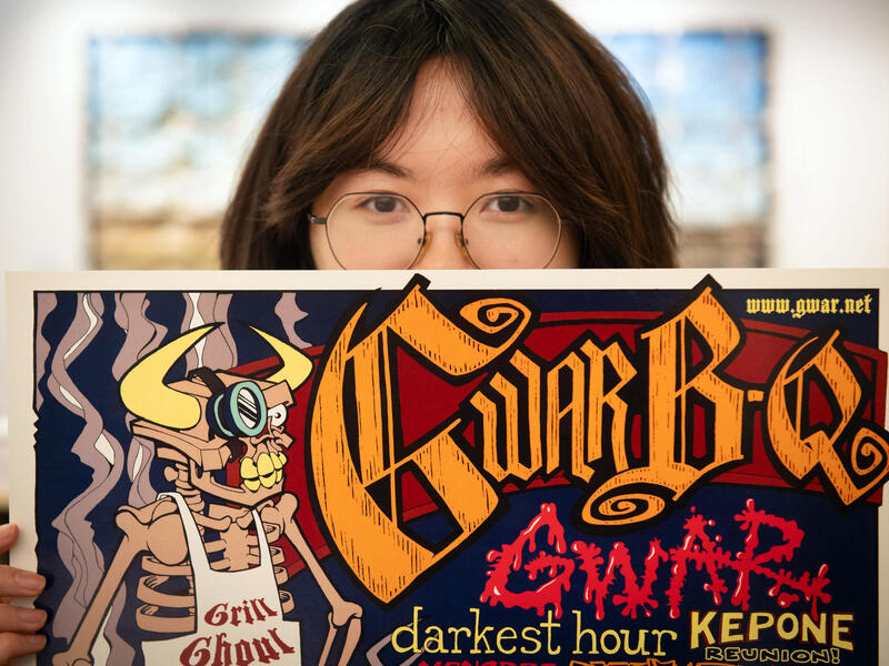 A person with glasses and long hair peers over the top of a poster.